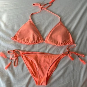 Korallrosa bikini från H&M med tofsar - Superfin korallrosa bikini från H&M med trekantsöverdel och knyt i både nacke och rygg. Underdelen har knyt i sidorna med dekorativa tofsar och små silvriga detaljer. Bikinin är i mjukt syntetmaterial och har diskret dekorationssöm längs kanterna. Perfekt för stranden i sommar. ÖVERDELEN ÄR STORLEK I 44 OCH UNDERDELEN I STORLEK 40