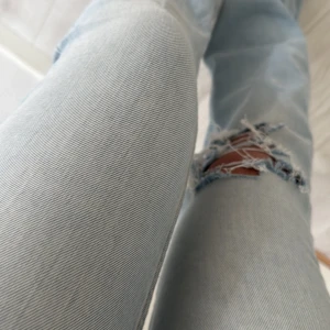 Ljusblå bootcut jeans med hål - Säljer ett par ljusblå jeans med bootcut-modell och ett stort slitet hål på ena knät. Jeansen har klassisk femficksdesign och är tillverkade i denim. Perfekta för dig som gillar en avslappnad och trendig look. Strl xs short length