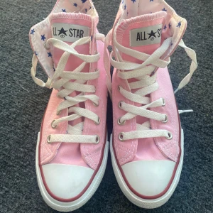 Rosa Converse All Star high-top sneakers - Snygga rosa Converse All Star high-top sneakers med vita snören och tåhätta. Insidan har ett mönster med blå stjärnor. Klassisk rund tå och platt sula, tillverkade i canvas med gummisula. Perfekta för dig som vill sticka ut med färg!