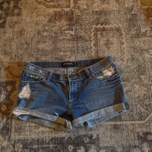 Lågmidjade jeansshorts  - Snygga blå jeansshorts från Abercrombie med slitna detaljer och upprullade benslut. Klassisk femficksmodell med låg midja och coola slitningar både fram och bak. Perfekta för varma dagar när du vill ha en avslappnad och trendig look.