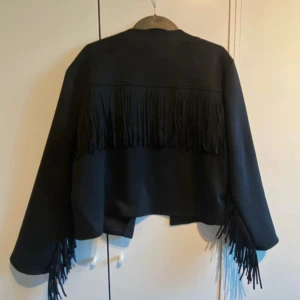 Svart jacka med fransar  - Tyvärr blev det fel med min beställning och jag kan inte skicka tillbaka jackan, så nu säljer jag den vidare. Jackan är helt ny och aldrig använd!  Nypris 700:-  🖤 Svart mockaimitation 🖤 Kraglös design & öppen framtill 🖤 Fransdetaljer på rygg och ärmar 🖤 Normal passform (regular fit)  Snygg och trendig jacka som passar perfekt till både vardag och fest.