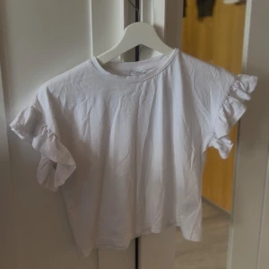 Vit volangtopp från Lindex - Supersöt vit topp från Lindex med volangdetaljer på ärmarna. Tillverkad i mjuk bomull och har en klassisk rund halsringning. Perfekt för dig som gillar en enkel men ändå lekfull stil.💞