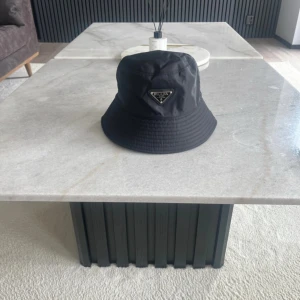 Svart bucket hat från Prada - Snygg svart bucket hat från Prada med klassisk siluett och ikonisk metallogo framtill. Hatten är tillverkad i ett lätt och slitstarkt syntetmaterial som ger en clean och modern look. Perfekt accessoar för att lyfta din outfit.