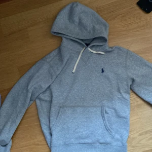 Grå hoodie från Polo Ralph Lauren - Snygg grå hoodie från Polo Ralph Lauren med klassisk blå logga broderad på bröstet. Tröjan har huva med vit snörning och en stor magficka. Tillverkad i mjuk bomullsmix, perfekt för chill dagar. Långärmad och normal passform.