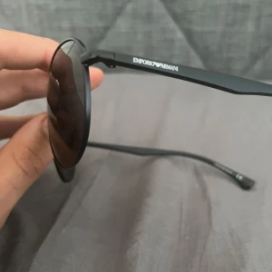 Svarta aviator-solglasögon Emporio Armani - Snygga svarta aviator-solglasögon från Emporio Armani med tunna metallbågar och mörka linser. Klassisk pilotform med dubbla bryggor och diskret logga på skalmen. Perfekt accessoar för en cool och stilren look. Ny skick utan repor eller defekter 