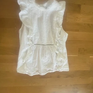 Vit volangblus från BikBok - Supersöt vit blus från BikBok med volangdetaljer längs axlarna och sidorna. Ärmlös modell med rynkade partier och dekorativa sömmar framtill. Perfekt för en fräsch och somrig look, lätt och luftig i bomull.