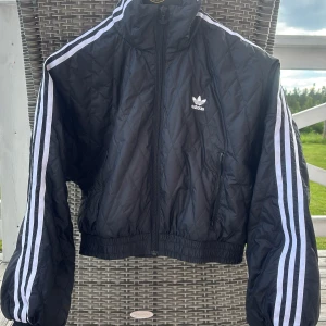 Svart quiltad jacka från Adidas - Quiltad svart jacka från Adidas med klassiska vita ränder längs ärmarna och broderad logga på bröstet. Jackan har dragkedja framtill, höga muddar och sportig vibe. Tillverkad i återvunnet Primegreen-material. Perfekt för dig som gillar streetstyle.