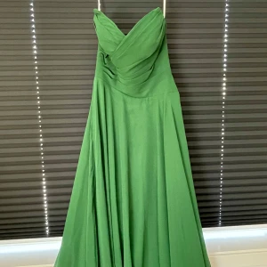 Grön axelbandslös långklänning - Snygg grön långklänning med axelbandslös design och draperad överdel. Klänningen har en vid, svepande kjol och är tillverkad i ett mjukt, lätt material som faller fint. Perfekt för dig som vill sticka ut med en elegant och stilren look.