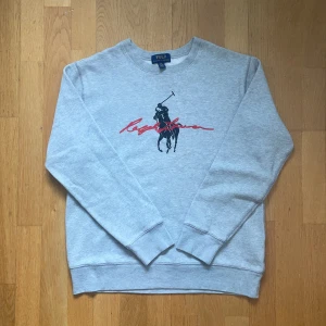 Grå sweatshirt från Polo Ralph Lauren - Säljer en klassisk grå sweatshirt från Polo Ralph Lauren med stort svart och rött tryck på bröstet. Tröjan har rund halsringning och långa ärmar. Perfekt för en avslappnad och stilren look.