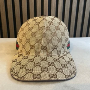Gucci keps med GG-mönster och ränder - Snygg keps från Gucci i beige och brunt med klassiskt GG-monogram över hela kepsen. På sidan finns de ikoniska gröna och röda ränderna. Justerbar rem baktill i brunt läder. Perfekt accessoar för dig som vill sticka ut med lyxig streetstyle.
