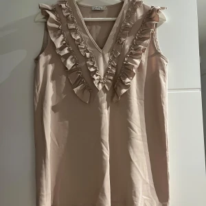 Beige ärmlös blus med volang - Supersöt beige blus från Simplicity i ärmlös modell med v-ringning och volangdetaljer framtill. Toppens material känns lätt och flowy, perfekt för en fräsch look. Volangerna ger en extra touch och gör toppen unik och feminin.