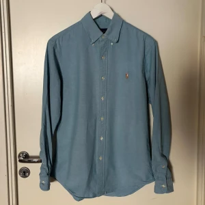 Blå Ralph Lauren skjorta - En blå skjorta från Ralph Lauren i storleken M och som är normal i storleken. Skjortan har ett mycket bra skick fast ett litet hål vid ena armen. Vid funderingar är det bara att höra av sig! 
