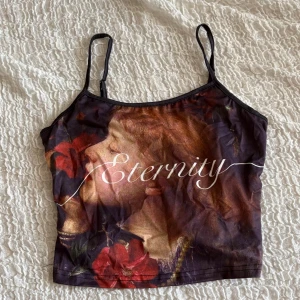 croppad linnetopp med text - Säljer en croppad linnetopp med smala, justerbara axelband och tryckt text framtill. Toppen har ett konstnärligt blommönster i mörka nyanser av rött, brunt och svart. Perfekt för dig som gillar unika prints och vill sticka ut.
