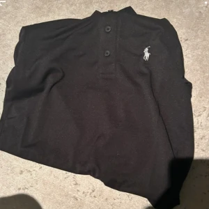 Svart pikétröja från Polo Ralph Lauren - Klassisk svart pikétröja från Polo Ralph Lauren med vit broderad logga på bröstet. Tröjan har korta ärmar, krage och tre knappar framtill. Tillverkad i mjuk bomull som känns skön mot huden. Perfekt för en clean och stilren look. Kommer med etikett.