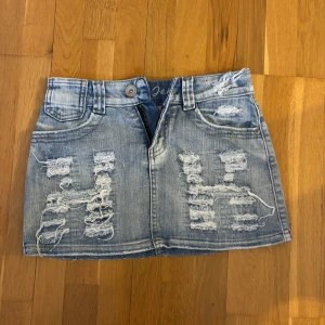 Blå jeanskjol - Snygg kort jeanskjol från Ping Jeans i ljusblå denim med slitna detaljer och råa kanter. Klassisk femficksmodell med dragkedja och knapp framtill. Perfekt för dig som gillar en edgy och trendig stil. Helt ny!!