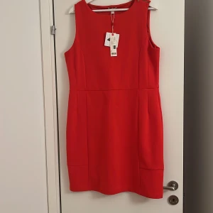 Röd ärmlös kortklänning - Snygg röd ärmlös klänning med rund halsringning och figurnära passform. Klänningen är kort och har stilrena sömdetaljer som ger en modern look. Perfekt för dig som vill sticka ut med en färgstark och enkel stil. Helt ny prislapp på har kostat 499kr.