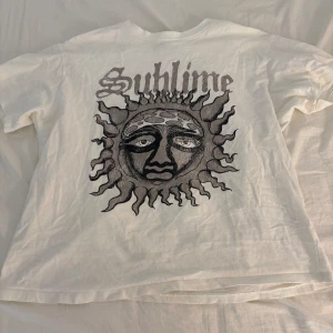 Vit Sublime t-shirt från H&M Divided  - Vit t-shirt från H&M Divided i samarbete med Sublime. Stor svart solgrafik och Sublime-tryck på ryggen. Klassisk passform med rund hals och korta ärmar. Tillverkad i mjuk bomull, perfekt för dig som gillar bandmerch och streetstyle.