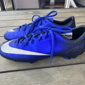 Blå Nike fotbollsskor med silverdetalj - Snygga blå fotbollsskor från Nike med stora silvriga swoosh-loggor på sidorna. Skorna har snörning och en smal, sportig form med svarta och blå detaljer vid hälen. Perfekta för dig som vill sticka ut på planen.