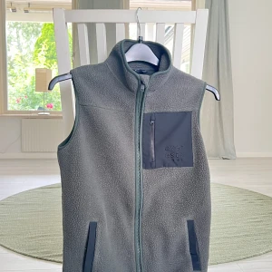 Grön fleeceväst Everest - Mysig grön fleeceväst från Everest med hög krage och hel dragkedja framtill. Västen har en svart bröstficka med dragkedja och två sidofickor. Perfekt för kyliga kvällar över en snygg skjorta.