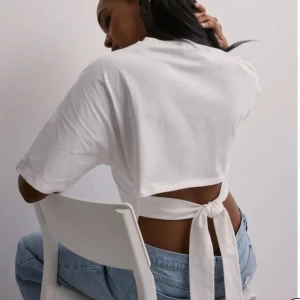 Vit croppad topp med knyt i ryggen - Säljer en vit croppad topp med kort ärm och snygg knytning i ryggen. Toppen har en enkel och clean look framtill, medan ryggen har öppen design med breda band som knyts till en rosett. Perfekt till jeans för en chill och trendig stil.