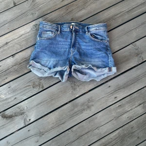 Blå jeansshorts från H&M, strl 38 - Snygga blå jeansshorts från H&M i klassisk modell med fem fickor, råa kanter och uppvikt benslut. Perfekta för sommaren och lätta att matcha med allt. Dem är värken hög elle lågmidjade utan är någon stans i mitten. 