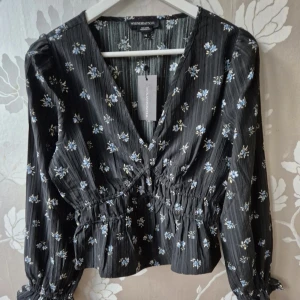 Svart blommig omlottblus från WEDNESDAYSGIRL - Supersöt svart omlottblus med små blå och vita blommor från WEDNESDAYSGIRL. Blusen har v-ringning, långa ärmar med resår vid ärmslut och smock i midjan för en snygg passform. Perfekt till jeans eller kjol för en trendig look.