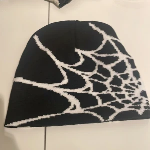 Y2K Mössa - Cool svart mössa med ett stort vitt spindelnätsmönster runt hela mössan. Perfekt accessoar för dig som vill sticka ut och addera lite edge till din outfit. Materialet är stickat och känns mjukt mot huvudet. Passar både till streetwear och mer alternativa stilar.