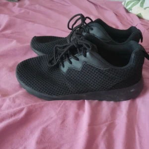 Svarta mesh-sneakers med snörning - Säljer ett par helsvarta sneakers med mesh-ovandel och snygga detaljer i syntet. Skorna har rund tå, platt sula och klassisk snörning. Perfekta för dig som gillar en clean och sportig look.