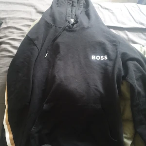 Svart hoodie från Hugo Boss med logga - Svart hoodie från Hugo Boss med vit BOSS-logga på bröstet. Tröjan har huva med snörning, känguruficka och en vit/guldig rand längs ärmarna. Tillverkad i mjuk bomull och polyester, perfekt för en chill och stilren look.