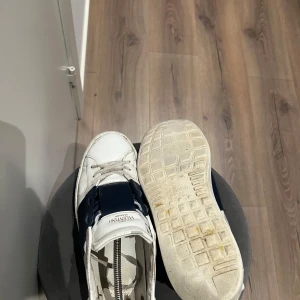Valentino Open Marinblå - Säljer ett par marin blå Valentino Open sneakers med bred mörkblå rem över mitten. Skorna har rund tå, snörning och klassisk platt sula. Ikoniska nitar baktill på sulan och diskret Valentino-logga på plösen. Medföljer box. Storlek: 46 