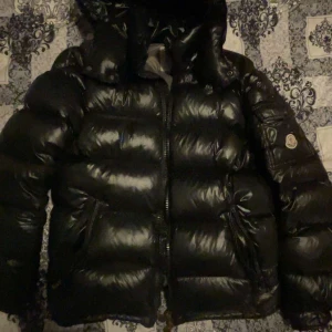 Svart Moncler jacka  - Säljer en svart, glansig dunjacka från Moncler med huva och dragkedja. Jackan har Moncler-logga på ärmen, flera fickor och klassisk pufferstil. Insidan har ett unikt serietidningsmärke. Perfekt för kalla dagar och riktigt snygg streetstil.
