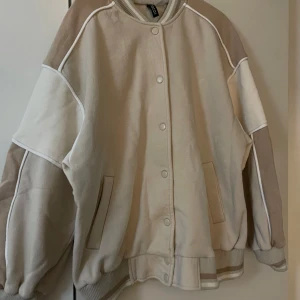 Beige bomberjacka från H&M, strl S - Säljer en oversized bomberjacka från H&M i olika beige toner. Jackan har ribbade muddar, tryckknappar framtill och coola kontrastpaneler på ärmarna. Perfekt för dig som gillar en avslappnad och trendig stil.