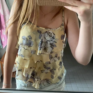 Blommig linnetopp från Guy Laroche - Supersöt linnetopp från Guy Laroche i ljusgul nyans med stora blommiga mönster i grått och blått. Tunt, luftigt material med volangiga lager och smala axelband. Ett hål under ett lager som inte syns vid användning. Sitter som storlek S