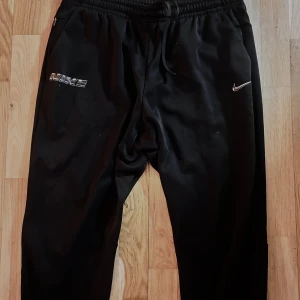 Nike Therma-Fit svarta mjukisbyxor - Svarta mjukisbyxor från Nike med Therma-Fit-teknologi. Byxorna har resår och snörning i midjan, samt silverfärgade Nike-loggor på båda sidor framtill. Perfekta för chill eller träning, riktigt sköna och stilrena.