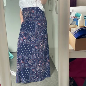Blå långkjol med paisleymönster - Supersnygg blå långkjol med patchwork-inspirerat paisleymönster i vitt och rött. Kjolen har hög midja och rak passform som faller snyggt längs benen. Perfekt för dig som gillar boho-vibe och vill sticka ut lite extra.