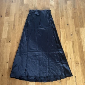 Svart satinkjol från Lager 157 - Snygg svart långkjol i satin från lager 157. Kjolen har en slät, glansig yta och stängs med dragkedja i sidan. 