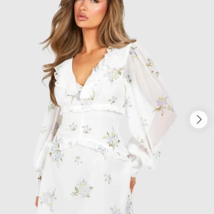 Vit blommig klänning med volang🌸 - Superfin klänning med blommigt mönster i pastellfärger. Klänningen har v-ringning, volangdetaljer och långa, transparenta ärmar. Perfekt för sommarens alla tillfällen. 