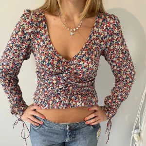 Blommig croppad blus från H&M - Supersöt blommig blus från H&M i småblommigt mönster med röda, blå och gula toner. Djup v-ringning fram, smockad rygg och långa ärmar med knyt vid handlederna. 💕Använt fåtal gånger❣️