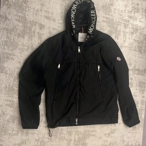 Svart vindjacka från Moncler med huva - Svart vindjacka från Moncler med stor huva och vit logotyptext längs kanten. Jackan har flera dragkedjefickor framtill, elastiska muddar och klassisk Moncler-logga på ärmen. Perfekt för dig som vill ha en stilren och sportig look.