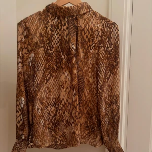 Brun skjorta med ormmönster - Säljer en snygg långärmad skjorta i brunt och beige med coolt ormmönster. Skjortan har en lös passform och är tillverkad i ett lätt, transparent material som ger en trendig vibe. Perfekt för dig som vill sticka ut med en unik look. Den har en knapp i ryggen också.