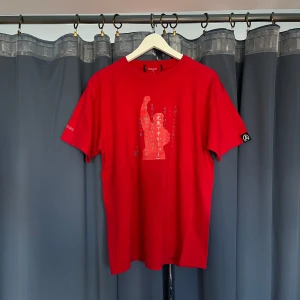 Röd t-shirt med tryck - Röd t-shirt med japansk text och grafiskt tryck framtill. Klassisk passform med rund hals och korta ärmar. Liten logga på ärmen och tryck på ryggen. Perfekt för dig som gillar streetwear med unik design. Köpt i Japan