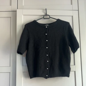 Svart kofta med pärlknappar - Kortärmad svart kofta med rund hals och dekorativa pärlknappar framtill. Tröjan har en mjuk stickad känsla och ribbade kanter vid ärmslut och nederkant. Perfekt för att ge en stilren touch till din outfit.