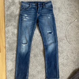 Slim jeans från Jack & Jones med slitnigar  - Säljer ett par helt nya  blå slim fit jeans från Jack & Jones, modell Slim/Glenn. Jeansen har slitna detaljer och hål på benen för en cool, avslappnad look. Klassisk femficksmodell med normal midja och smal passform. Perfekta till sneakers och hoodie.