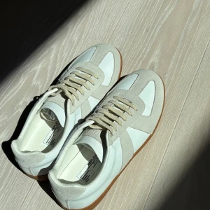 Maison margiela - Snygga vita sneakers med beige mockadetaljer och gummisula. Skorna har klassisk design med rund tå och snörning. Perfekta för dig som gillar stilrena och minimalistiska vibes. Materialmix av skinn och mocka ger en lyxig känsla. Har inget kvitto. Pris går att diskuteras