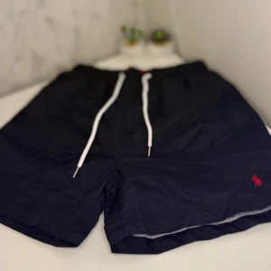 Polo Ralph Lauren Badshorts - Svarta badshorts från Polo Ralph Lauren med vit snörning i midjan och röd broderad logga på benet. Shortsen har elastisk midja, innerbyxa i mesh och bakficka med lock. Perfekta för stranden eller poolen.