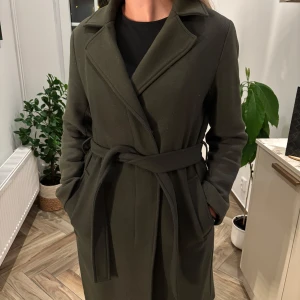 Olivgrön kappa från Filippa K, S - Snygg olivgrön kappa från Filippa K i klassisk modell med bälte i midjan och knappar framtill. Jackan har långa ärmar, bred krage och två fickor. Perfekt för dig som gillar stilrena och tidlösa plagg. Lite nopprig samt hårig pga hund men det löser vi fram till nån beställer.