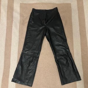 Svarta skinnbyxor från Sandro Paris - Svarta skinnbyxor från Sandro Paris i äkta läder. Byxorna har raka ben med lätt bootcut, hög midja och två bakfickor. Snygg och tidlös modell som ger en edgy vibe till din outfit. Perfekta att styla med en oversized hoodie eller en tight topp.