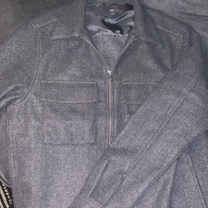 Grå overshirt från H&M med dragkedja - Snygg grå overshirt från H&M med dragkedja framtill och två stora bröstfickor. Jackan har en stilren look och är tillverkad i ett mjukt material som känns skönt mot huden. Perfekt att slänga på sig över en hoodie eller t-shirt.