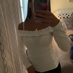 Vit offshoulder topp med volang - Säljer en vit offshoulder topp med lång ärm och snygg volang runt hela halsringningen. Toppen är figurnära och har en stilren, enkel look som passar perfekt till jeans eller kjol. Materialet känns mjukt och stretchigt, vilket gör den bekväm att bära.