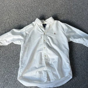 Vit skjorta från Ralph Lauren - Klassisk vit skjorta från Ralph Lauren med knappar framtill och broderad logga på bröstet. Skjortan har lång ärm, krage och är tillverkad i mjuk bomull. Perfekt för en clean och stilren look. Storleken är 16 y men passar xs i vuxen 